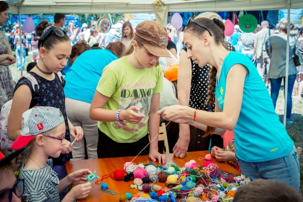 Several kids and a woman around a table filled with balls of yarn at a fair.