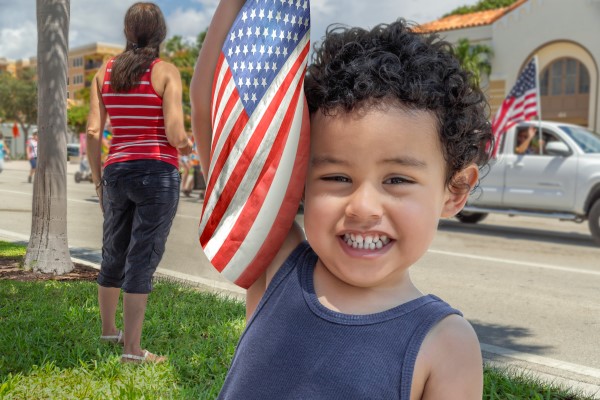 A little boy with curly hair smiling and holding an American flag.