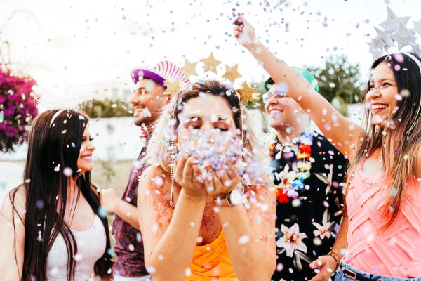 A woman at an outdoor celebration blowing confetti at the camera. She is surrounded by other men and women at the festivity.