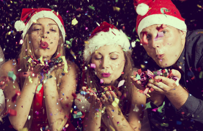 Two women and a man in Santa hats blowing confetti at the camera.