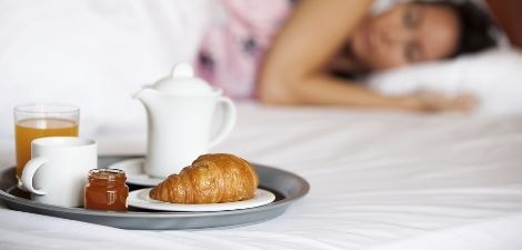 Pastry, coffee, and juice on a breakfast platter on the bed next to a sleeping woman.