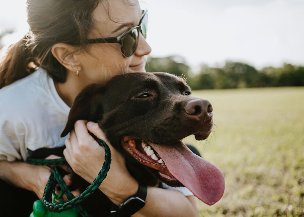 A woman in sunglasses hugging a panting dog outside.