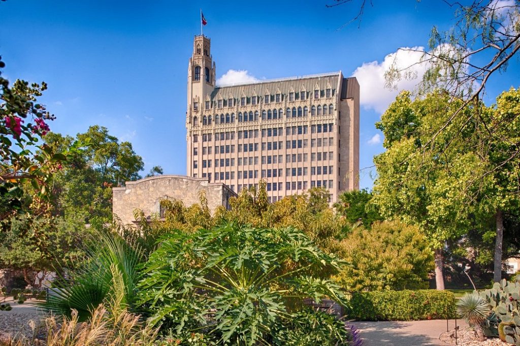 View of the Emily Morgan Hotel with the top of the Alamo in the foreground amidst some trees.