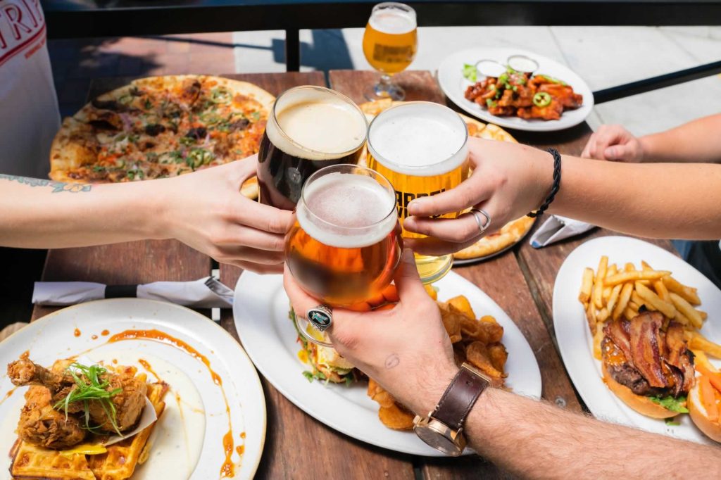Friends raising glasses of beer over plates of comfort food.