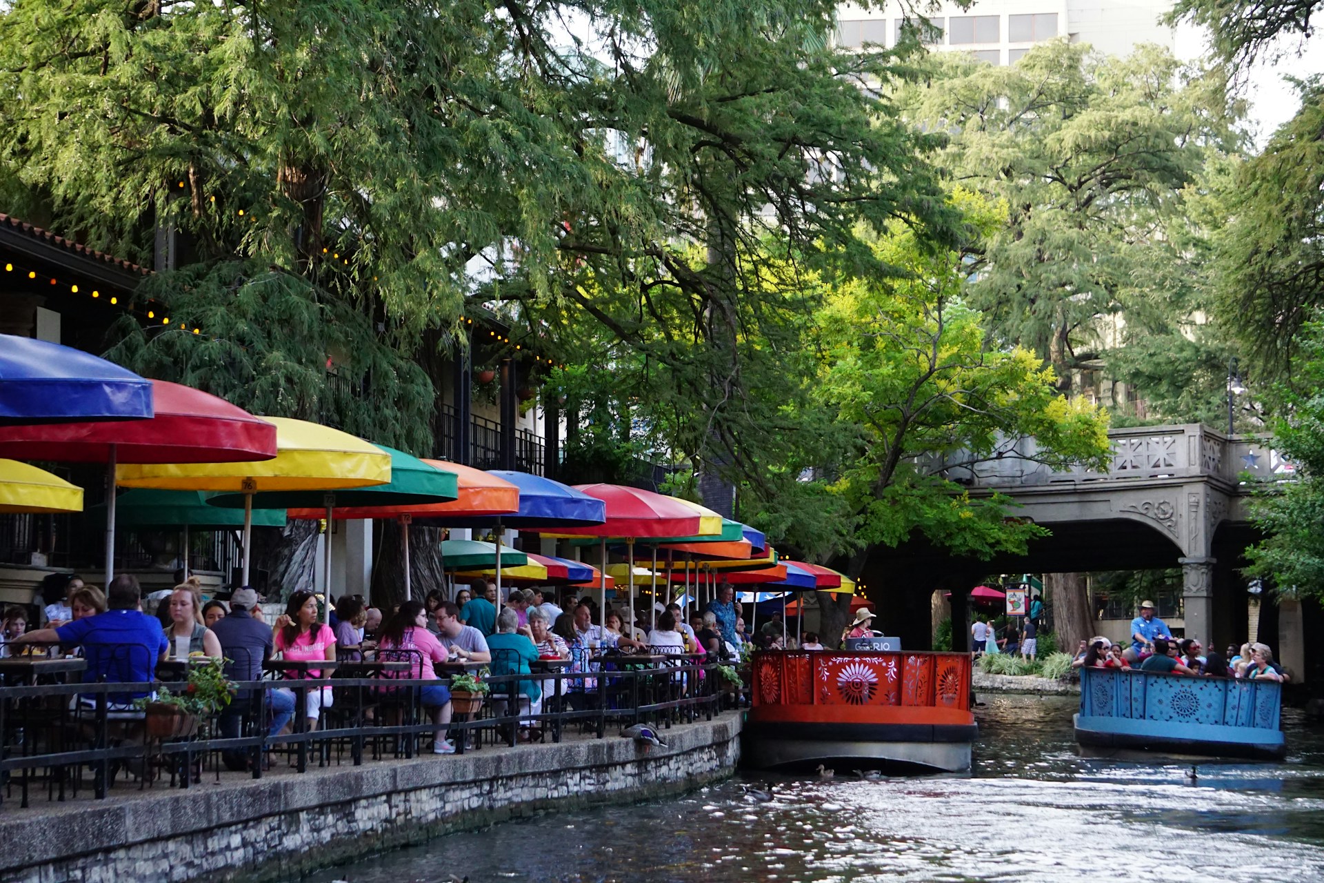 People dining at restaurants along the San Antonio River Walk while boats transport tourists down the river.