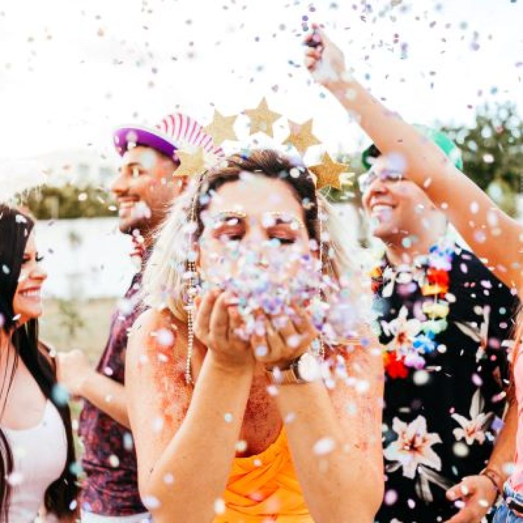 A woman at an outdoor celebration blowing confetti at the camera. She is surrounded by other men and women at the festivity.